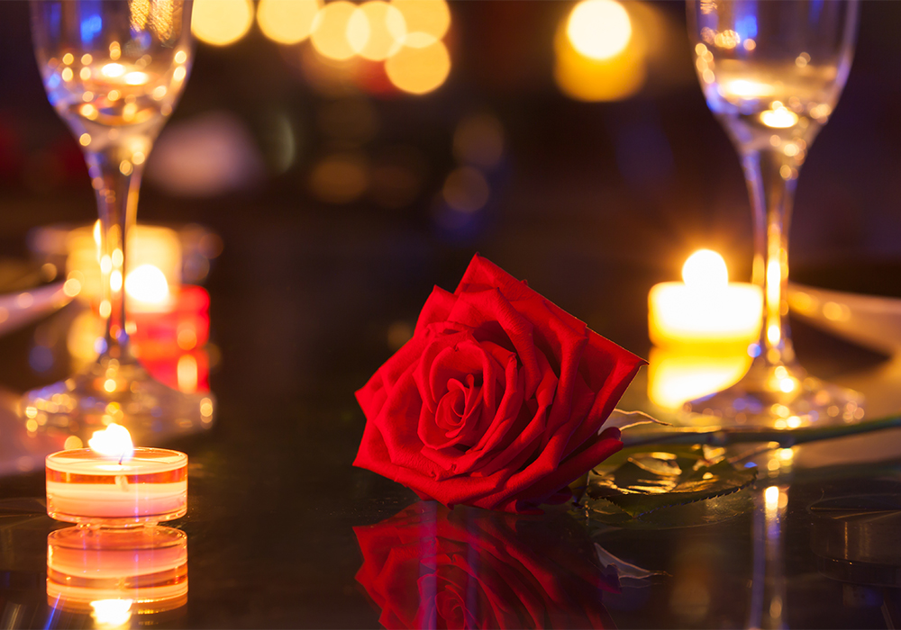 レストランテーブルの上でキャンドルのあたたかい灯りに照らされる一輪の赤い薔薇